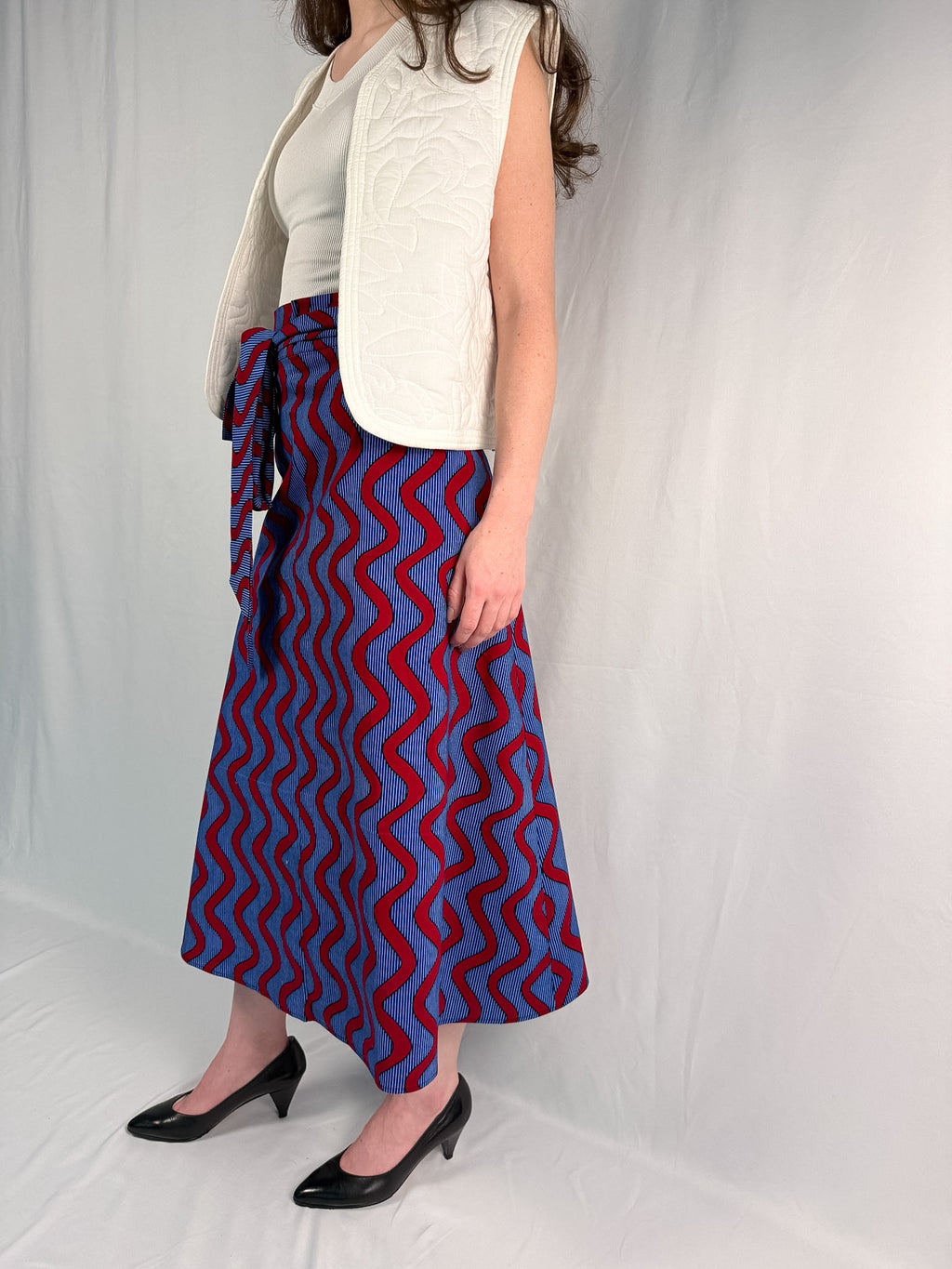 Person wearing a blue and red patterned skirt with a white top and vest on a plain background - onsem