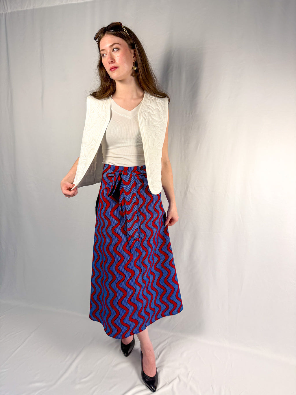 Woman wearing a white top, cream cardigan, and blue and red patterned skirt against a plain background - onsem